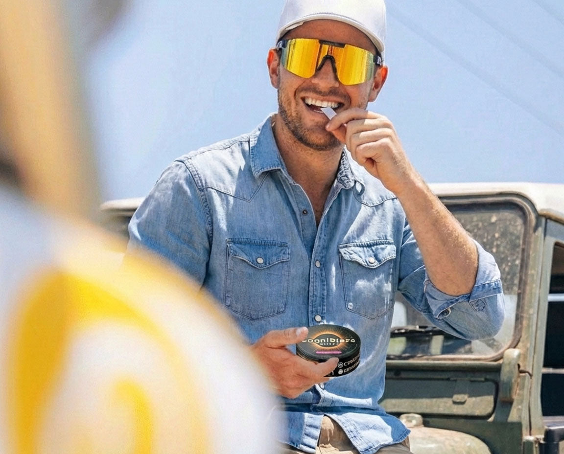Man wearing sunglasses and a cap, sitting in front of a vehicle, holding a can of energy pouches.