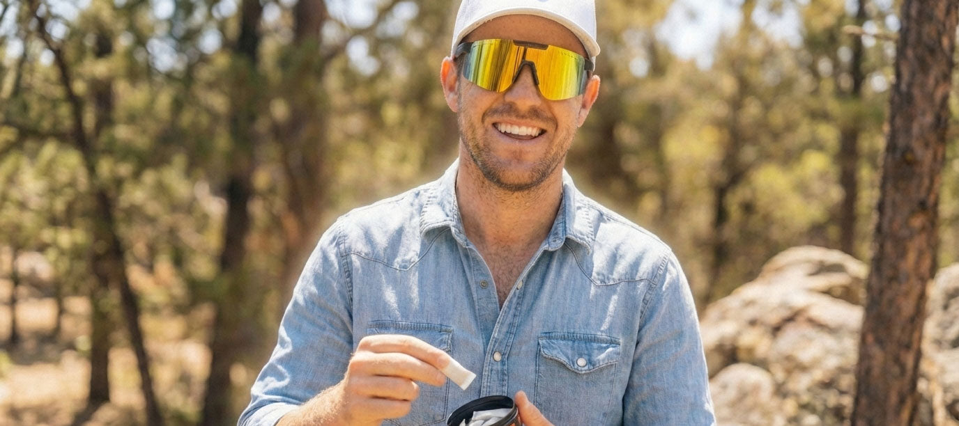 Man wearing sunglasses and a cap outdoors with trees in the background, holding a can of nootropic pouches